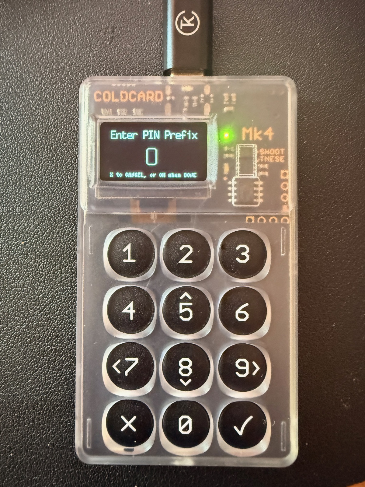 The Coldcard Mk4 hardware wallet mid-PIN-entry. The transparent case reveals the circuit board and secure element chip. A small green status LED indicates the device is powered. The OLED screen displays 'Enter PIN Prefix' with a numeric counter. A physical 12-button keypad is below the screen.