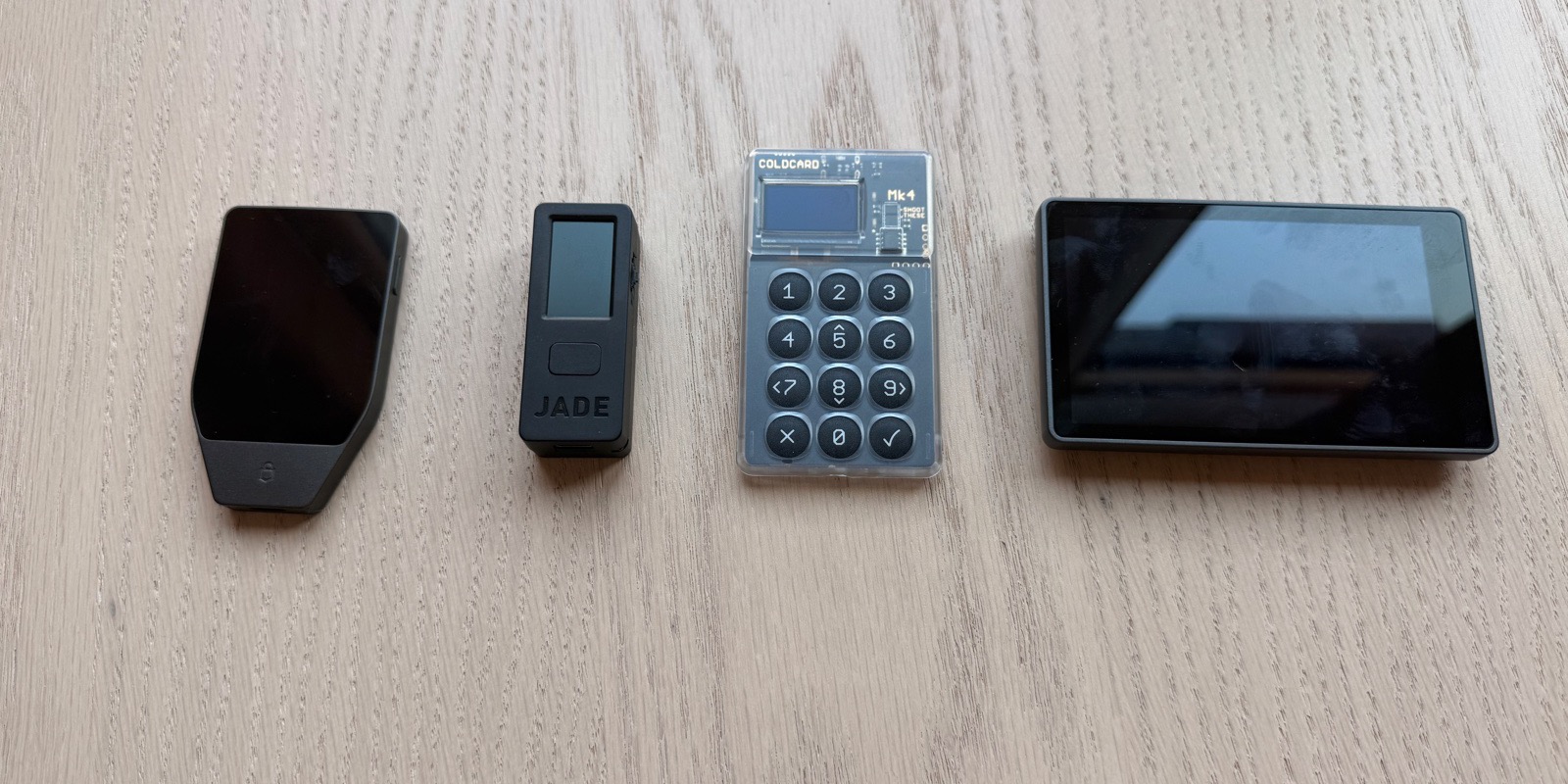 Four Bitcoin hardware wallets from four different vendors on a light wood surface, left to right: Trezor Safe 5 (vertical touchscreen with secure element), Blockstream Jade (compact single-button stick), Coldcard Mk4 (transparent case showing the circuit board and physical numeric keypad), and Keystone 3 Pro (large landscape touchscreen with fingerprint sensor and QR-only air-gapped operation). Four different design philosophies for the same problem: keeping a private key off an internet-connected device.