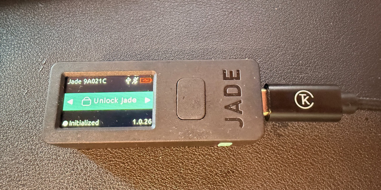 The Blockstream Jade hardware wallet powered on, displaying its unlock screen. The compact device shows 'Unlock Jade' on a small color screen, with status indicators for initialization and firmware version. The JADE wordmark is embossed on the side.