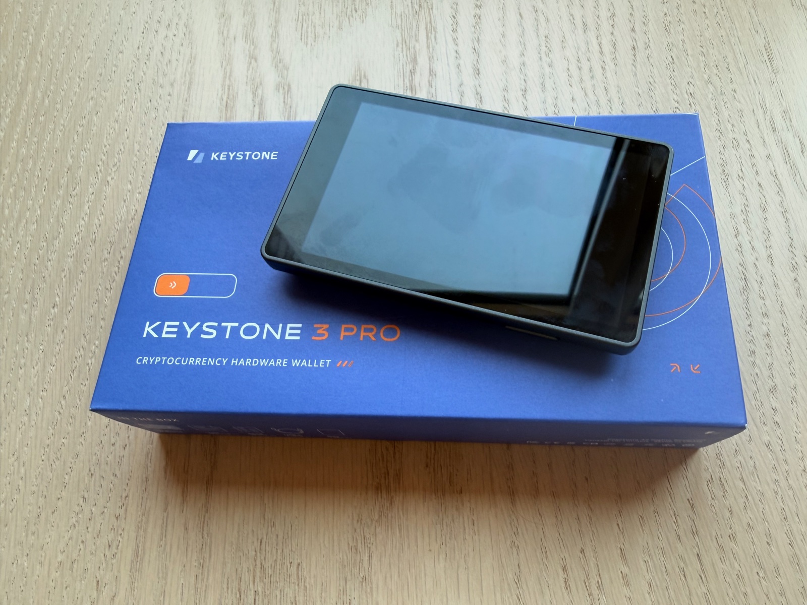 The Keystone 3 Pro hardware wallet resting on its blue retail packaging. The device's landscape touchscreen is dark; the box shows the Keystone wordmark and product name. Light wood surface in the background.