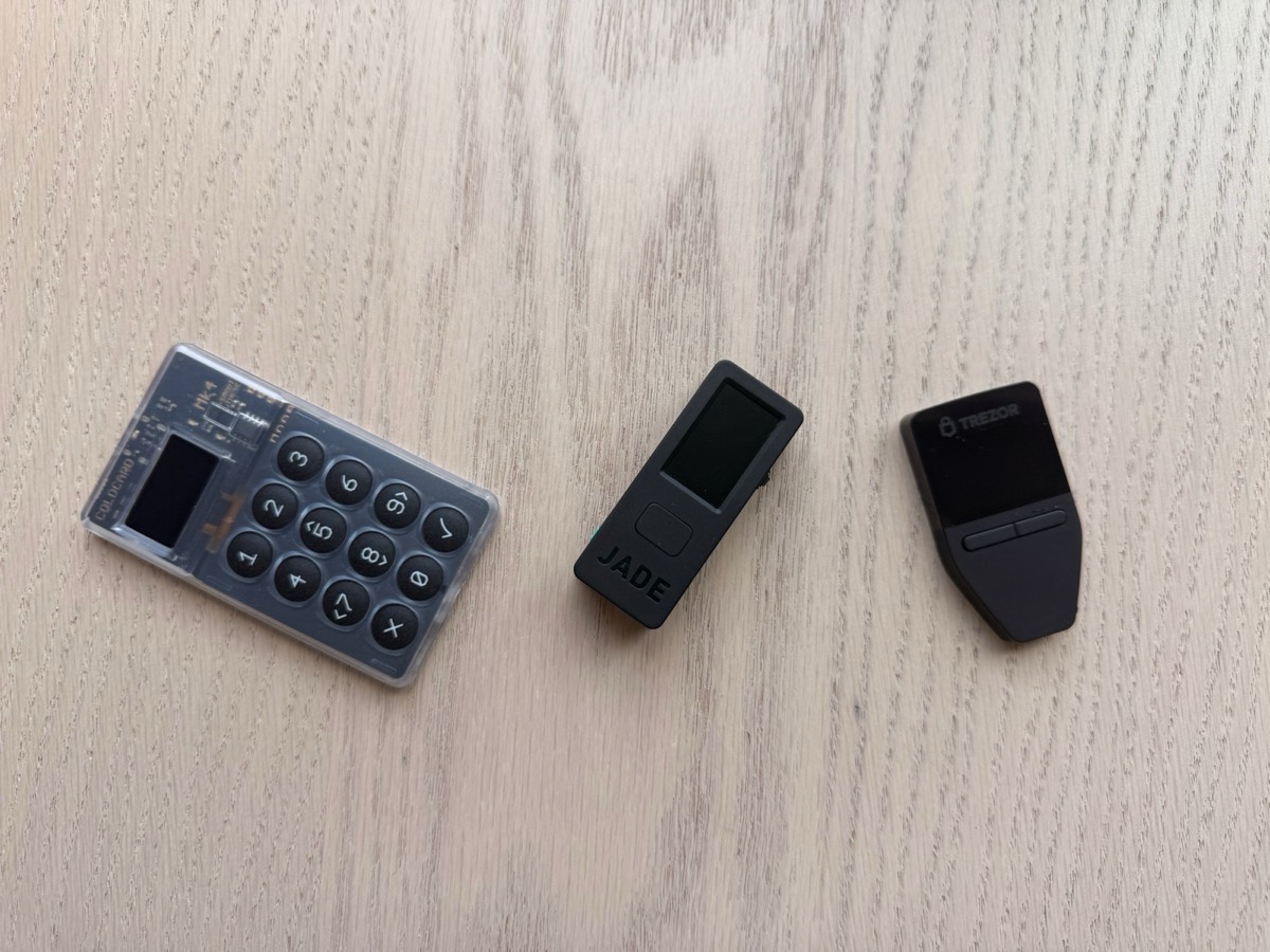 Three hardware wallets in a row on light wood: a Coldcard Mk4 keypad device on the left, a Blockstream Jade in the middle, and a Trezor Safe on the right.