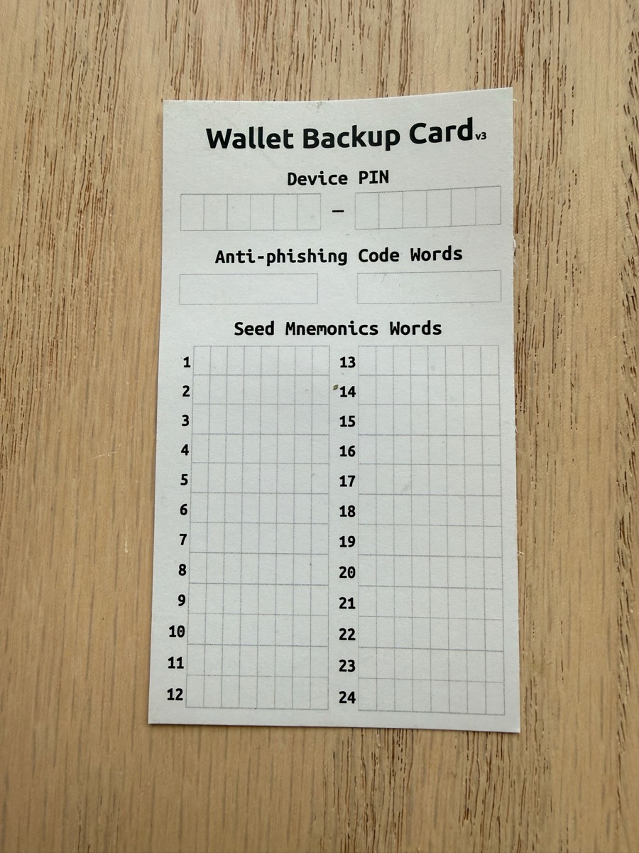A blank Trezor paper recovery card on light wood, printed with sections for device PIN, anti-phishing code words, and a 24-word seed grid.
