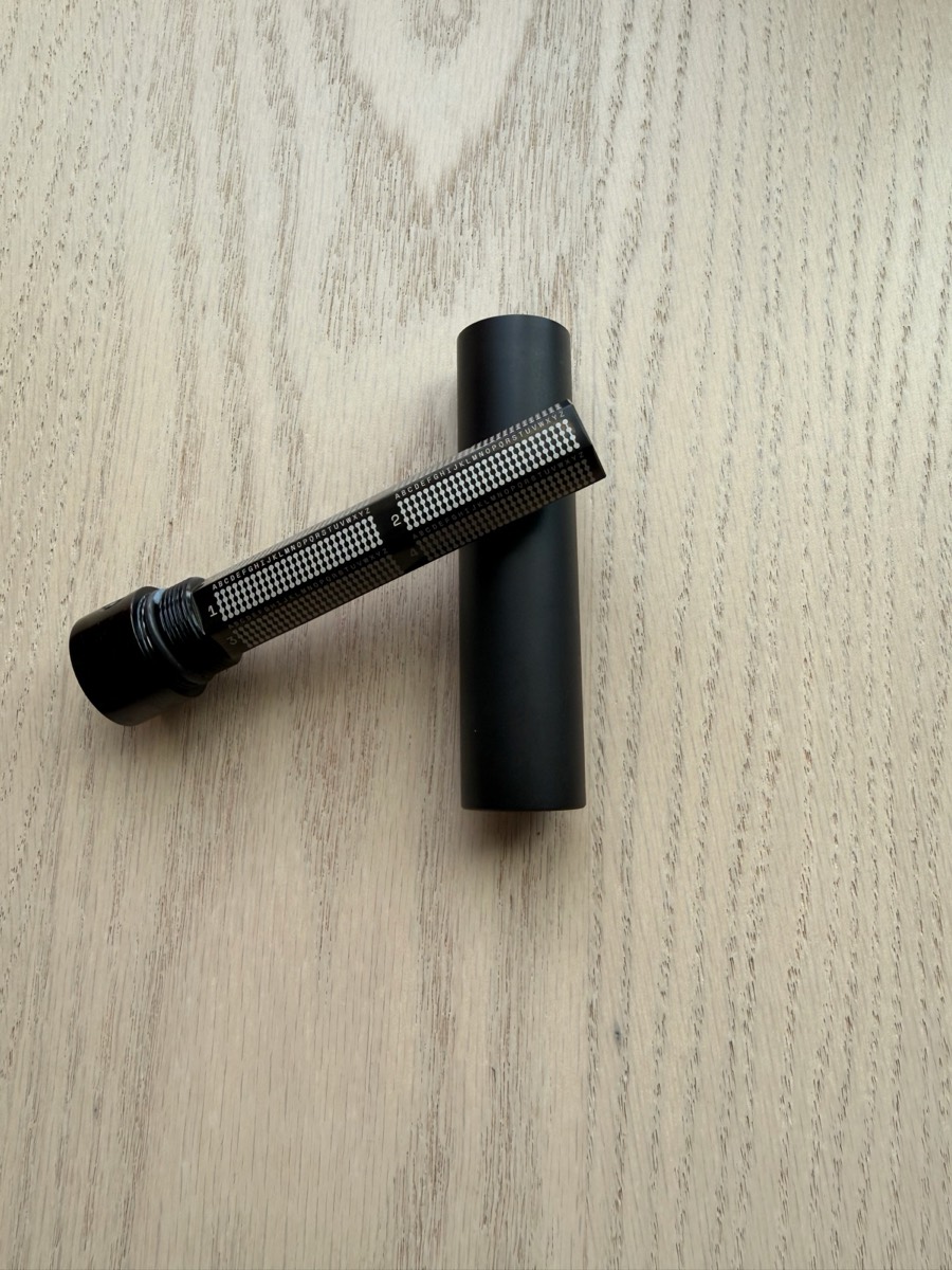 A tube-style stainless-steel seed backup on light wood with the protective sleeve removed. Each letter is marked by punching dots into a fixed grid. The shaft is blank.