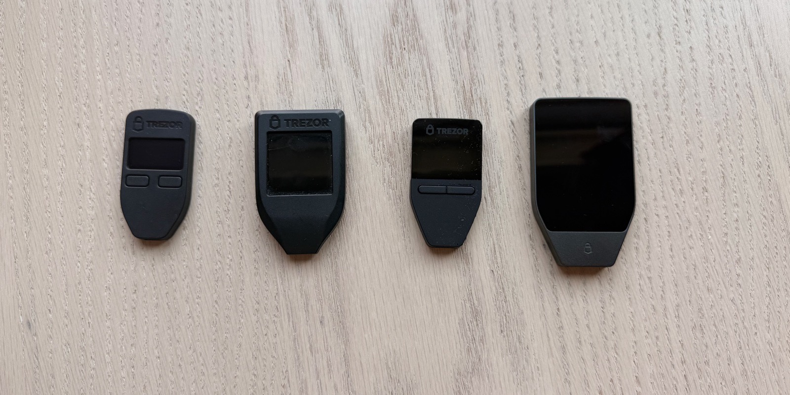 Four Trezor hardware wallets in chronological order on a light wood surface, left to right: Trezor One (2014), Trezor Model T (2018), Trezor Safe 3 (2023), Trezor Safe 5 (2024). The shapes and screen sizes show a decade of progression - from the original two-button monochrome design to the current touchscreen flagship with a secure element.