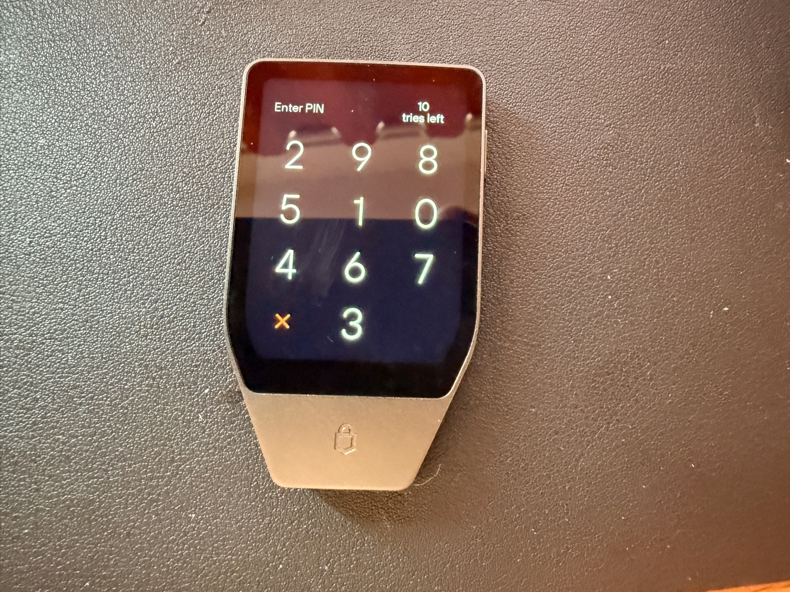 The Trezor Safe 5 hardware wallet powered on, displaying its scrambled PIN entry screen. The number positions are randomized on each unlock to prevent shoulder-surfing and smudge-pattern attacks. The device sits on dark leather, with the embossed Trezor lock icon visible below the screen.